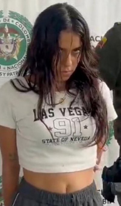 A woman with long dark hair, wearing a white cropped t-shirt that reads 'LAS VEGAS 91 STATE OF NEVADA', stands in front of a…