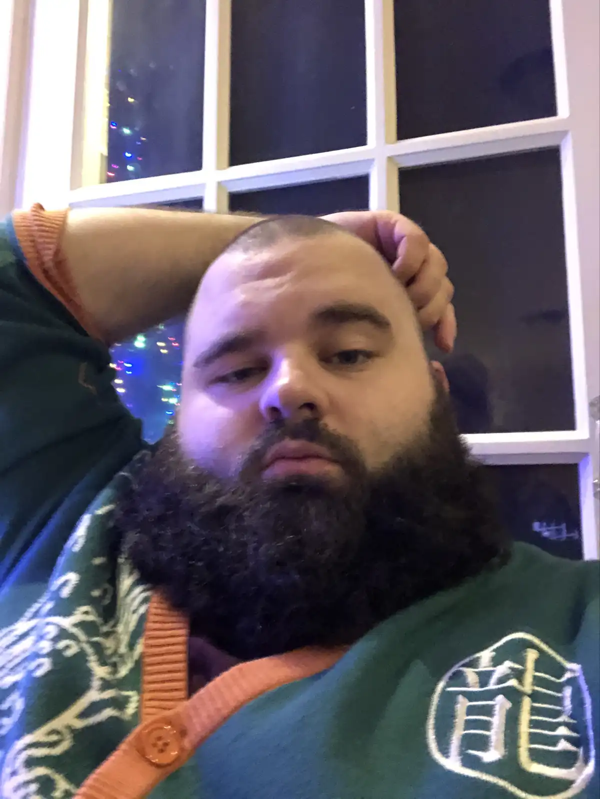 A man with a full beard and shaved head, wearing a green sweater with an orange collar, rests his head against a window with…