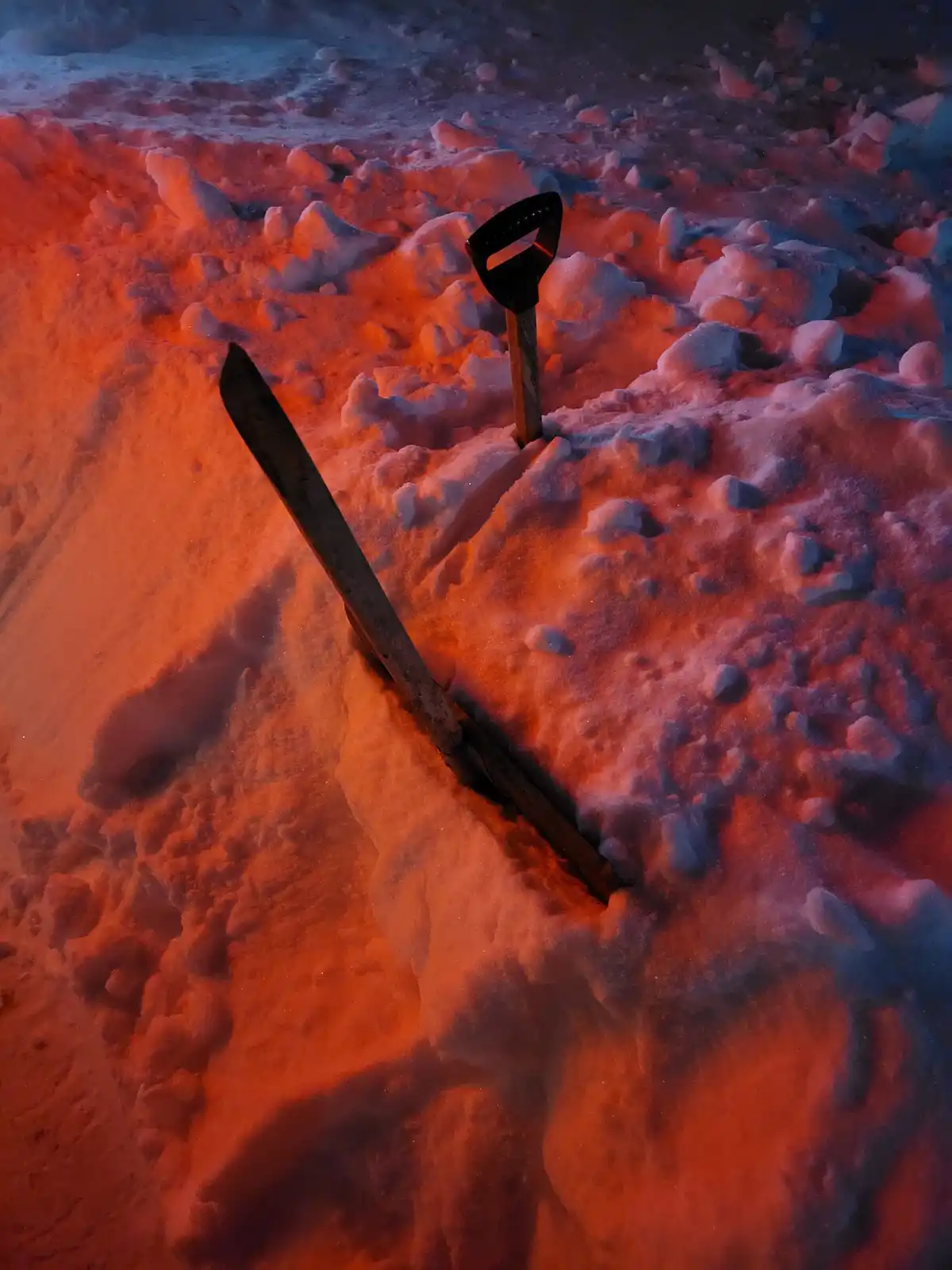 A snow shovel and a snow rake are stuck in a pile of snow under dramatic red and blue lighting. The overlaid text reads…