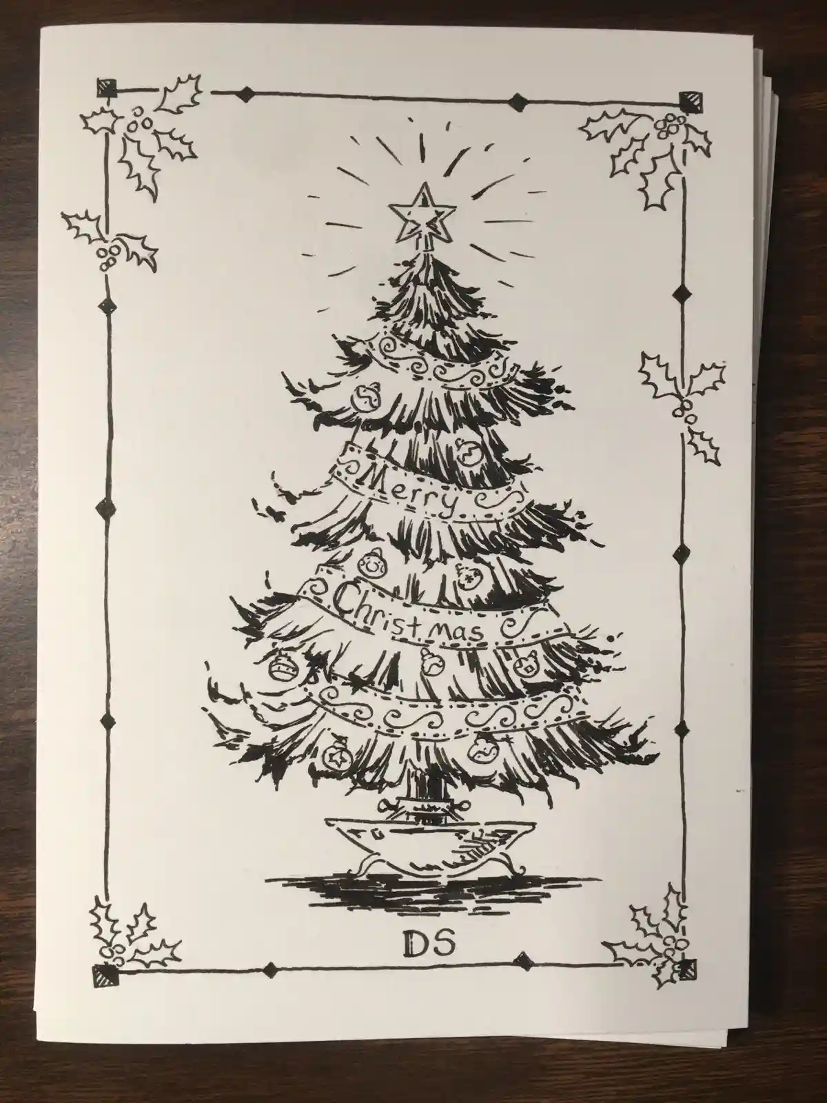 A hand-drawn black and white Christmas card features a decorated tree with a star on top, framed by holly leaves. The tree…