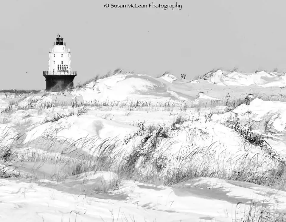 A black-and-white photograph shows a lighthouse standing on a dune-covered beach, with snow blanketing the sand and grasses…