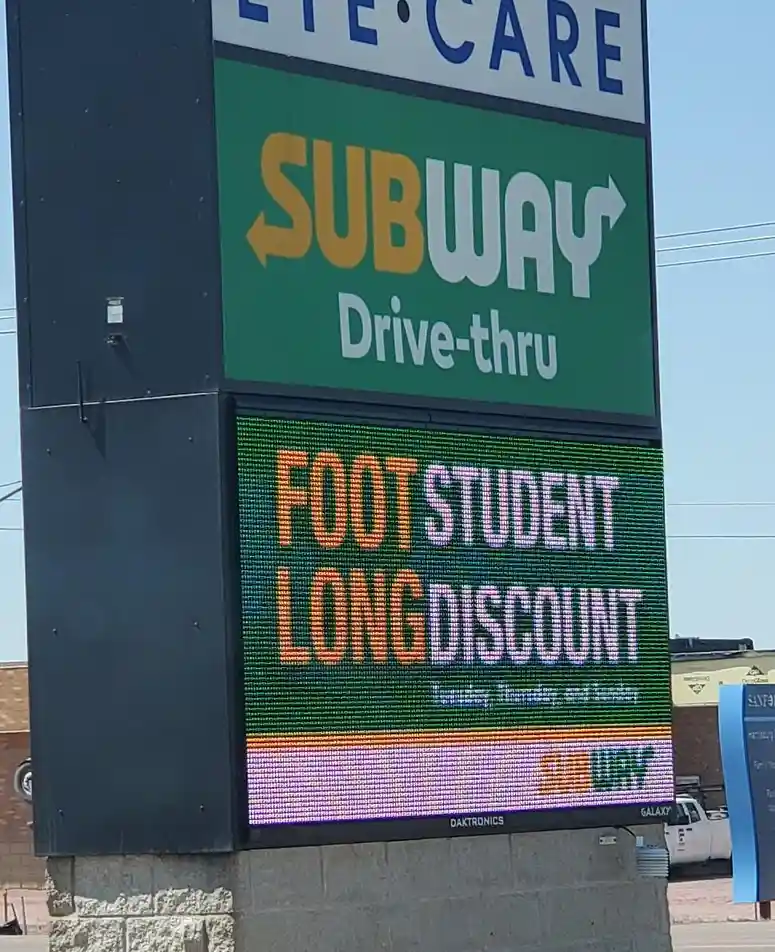 A tall outdoor sign for a Subway restaurant with a drive-thru, featuring a digital LED display below that shows the text…