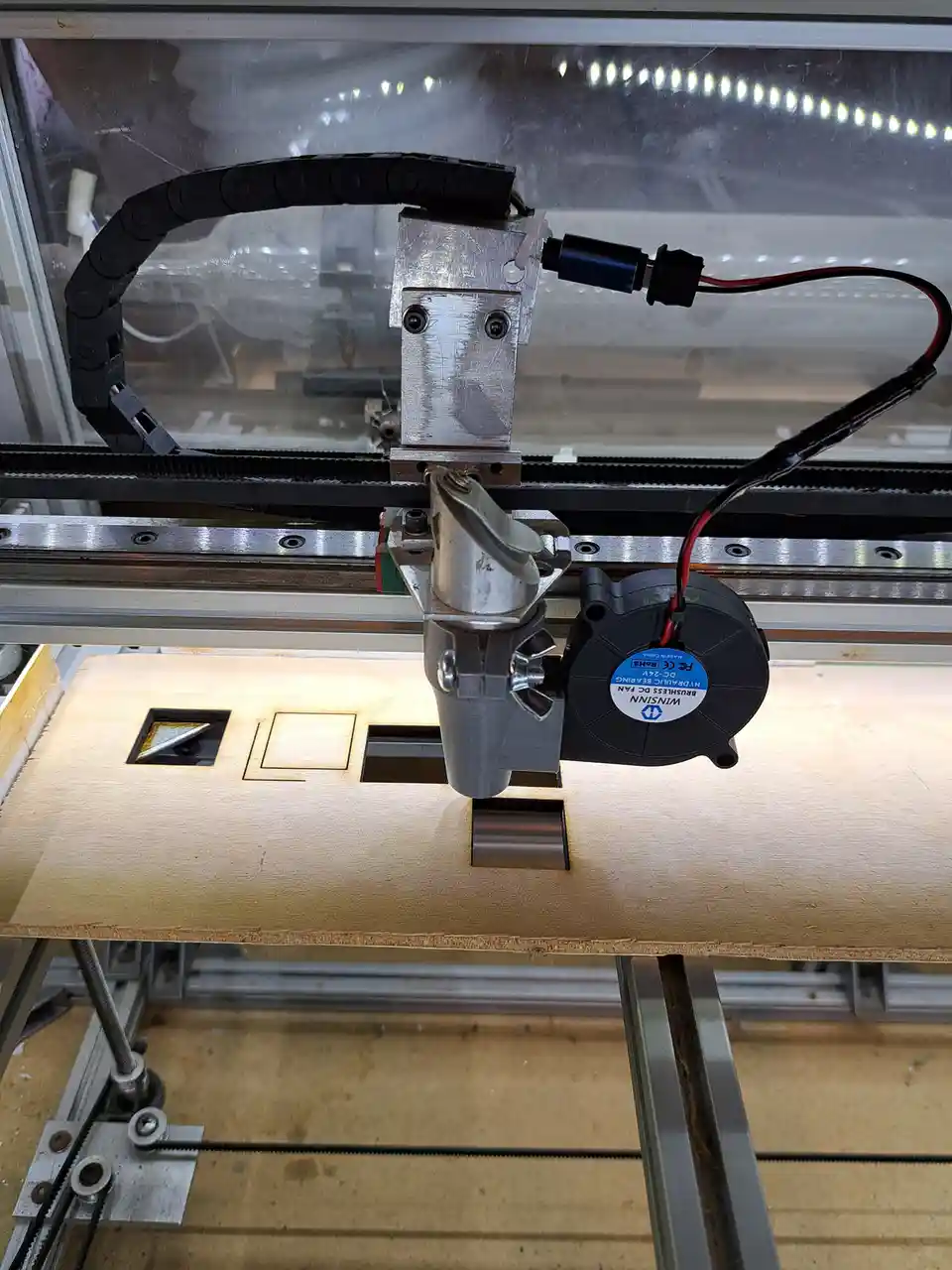 A close-up photo of a laser cutter's cutting head, which has a small black fan attached to it via wires and a bracket. The…