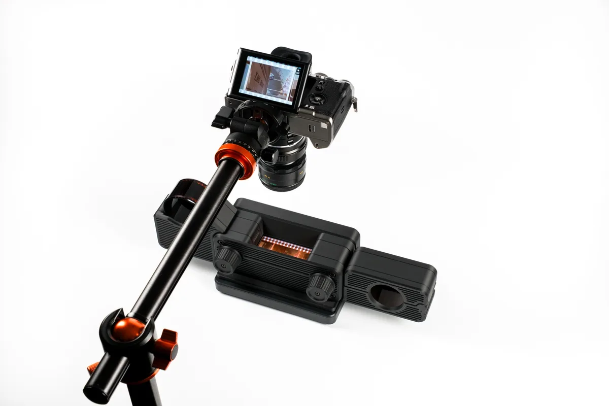 A digital camera mounted on a tripod arm is pointed at a black film scanner with a strip of film visible inside. The…