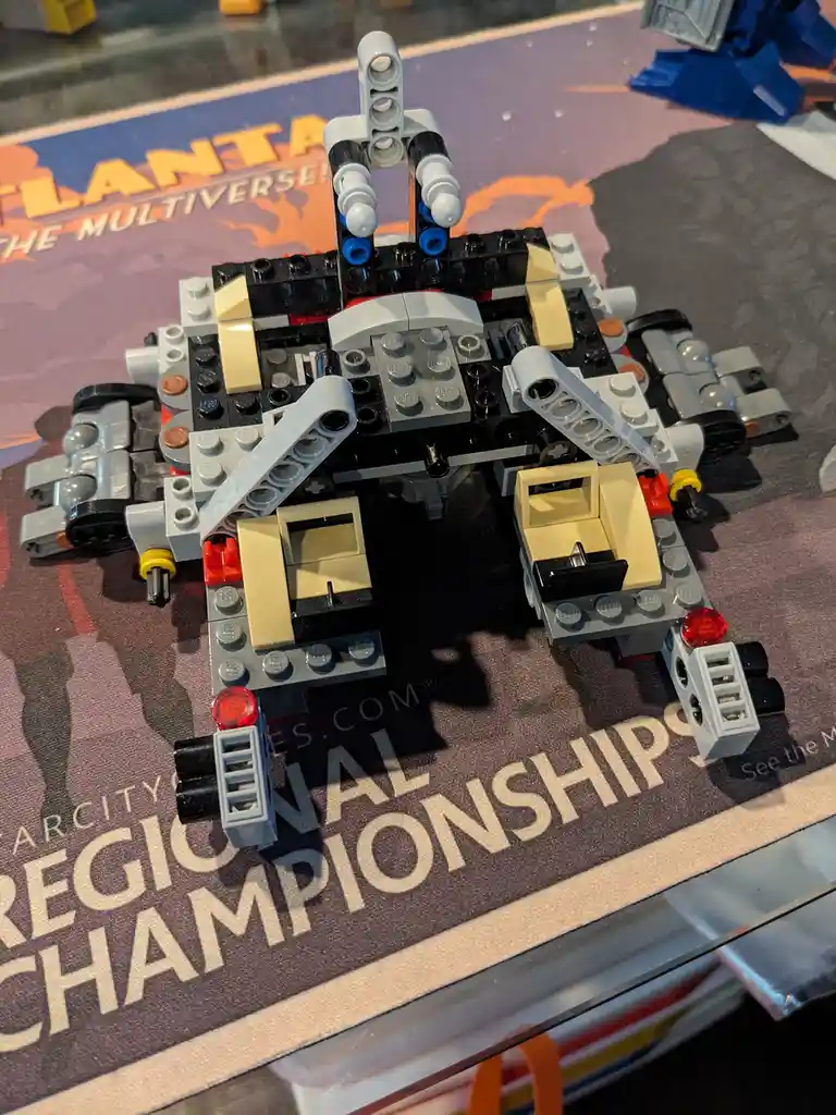 A close-up photo shows a complex LEGO robot built from black, gray, beige, and red pieces, sitting on a mat that reads…