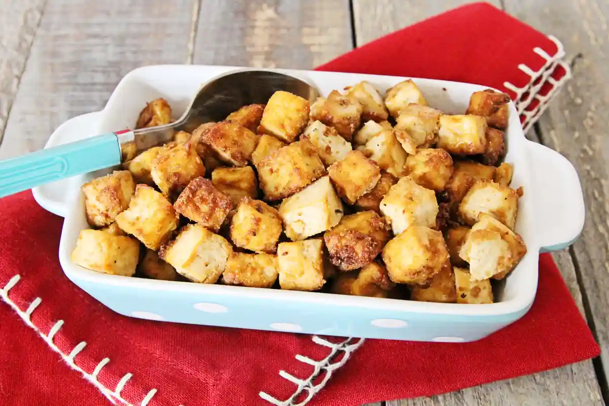 A white ceramic baking dish with light blue polka dots is filled with golden-brown, cubed tofu, resting on a red cloth with…