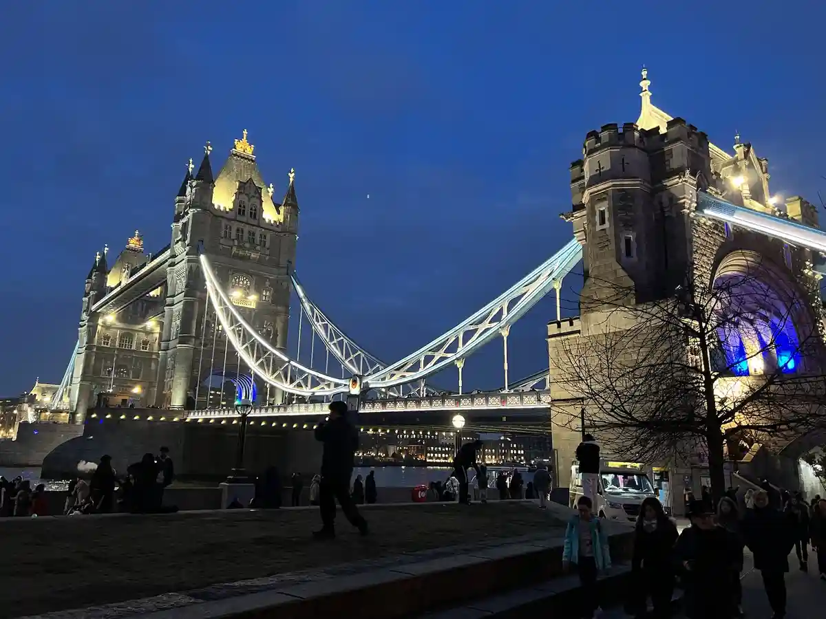 A nighttime photo of Tower Bridge in London, illuminated with blue and white lights against a dark blue sky. People are…