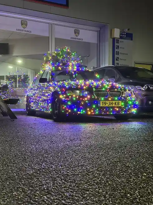 A dark-colored BMW sedan is parked outside a garage labeled 'BCZ ASSISTANCE' at night, completely covered in multicolored…
