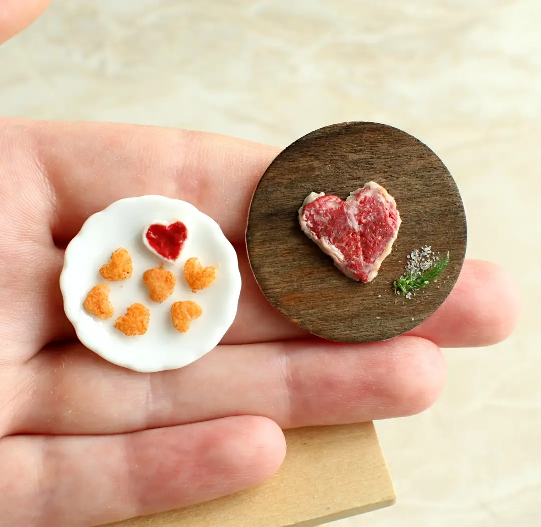 A hand holds two tiny, heart-themed food items: one is a white plate with heart-shaped cereal and a red heart-shaped jelly…