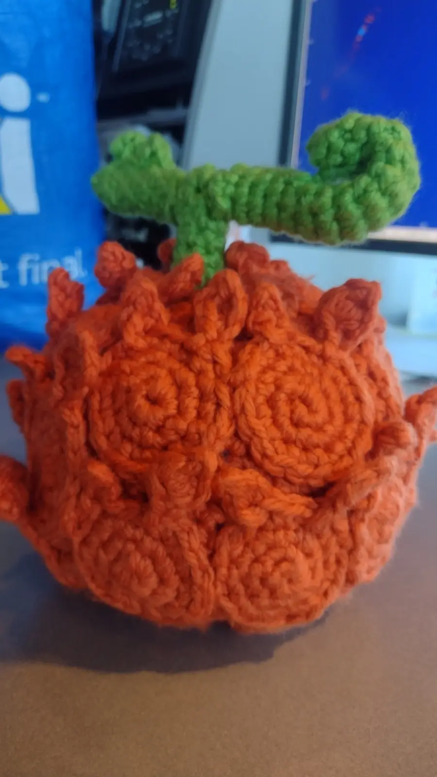 A close-up photo of a handmade, orange crocheted pumpkin with a green crocheted stem, sitting on a desk. The pumpkin has a…