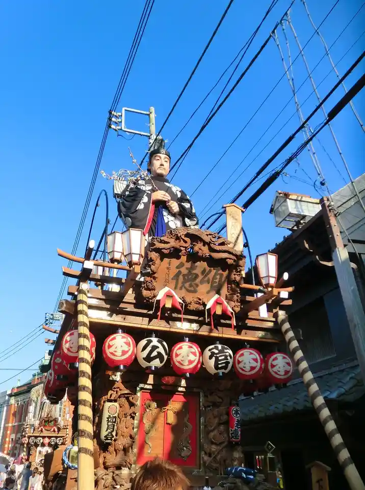 A low-angle photo shows a large, ornate Japanese festival float under a clear blue sky, with a statue of a man in…
