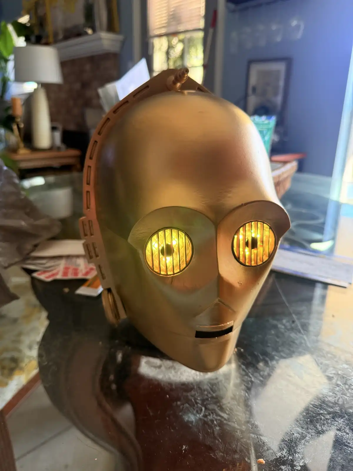 A golden C-3PO robot head from Star Wars sits on a glass table in a home, its eyes glowing yellow. The overlaid text reads…