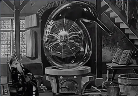 A black-and-white still from a vintage film shows a man in a lab coat sitting in a chair, looking up at a large glass sphere…