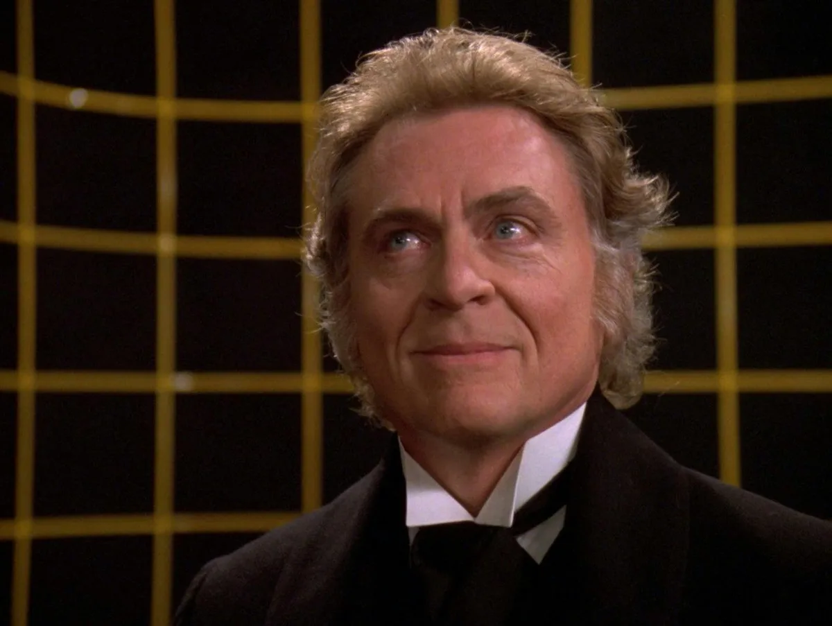 a man with blond hair and blue eyes, wearing a black suit and bowtie, looks upward with a knowing, slightly smug expression…