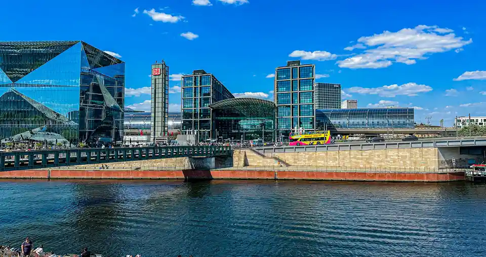 A bright, sunny photo of Berlin Hauptbahnhof with its distinctive glass architecture and a river in the foreground. A yellow…