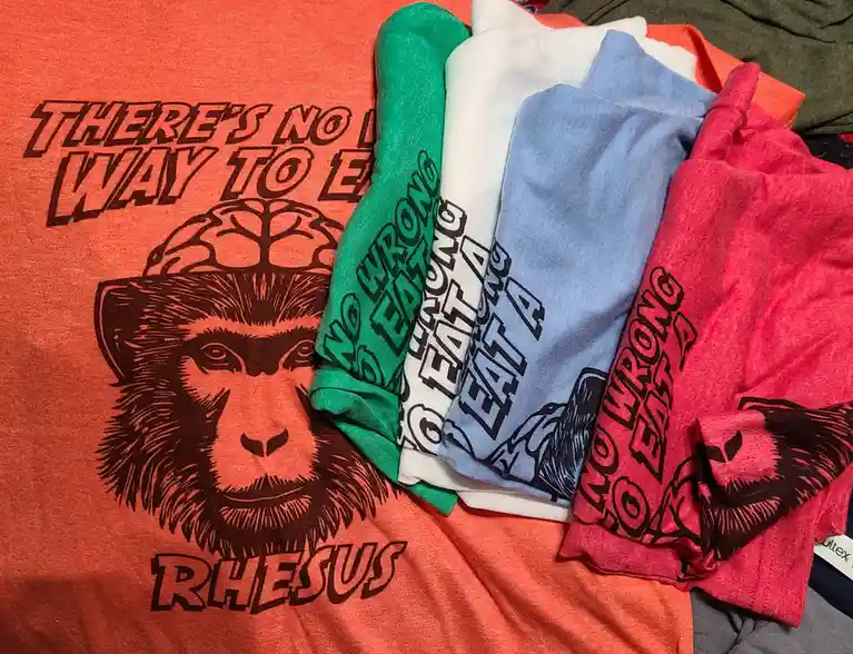 A pile of colorful t-shirts, with the largest orange one in the foreground featuring a graphic of a monkey wearing a brain…
