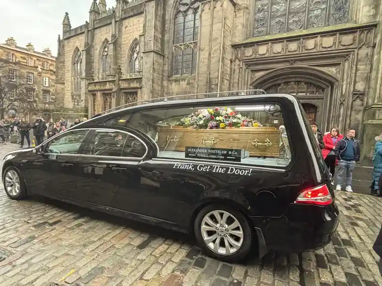 A black hearse is parked on a cobblestone street in front of a Gothic-style stone church, with a wooden coffin visible…