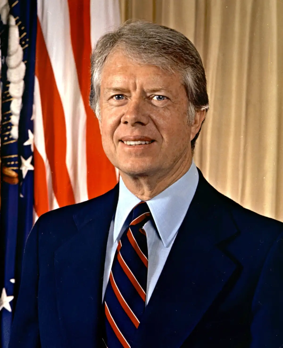 A formal portrait of former U.S. President Jimmy Carter, smiling, wearing a dark blue suit, light blue shirt, and a striped…