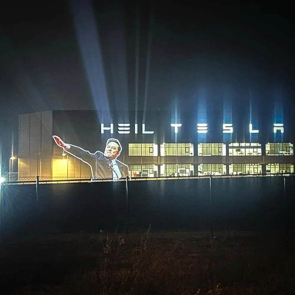 A nighttime photo of a large industrial building with bright spotlights shining upwards. Projected onto the side of the…