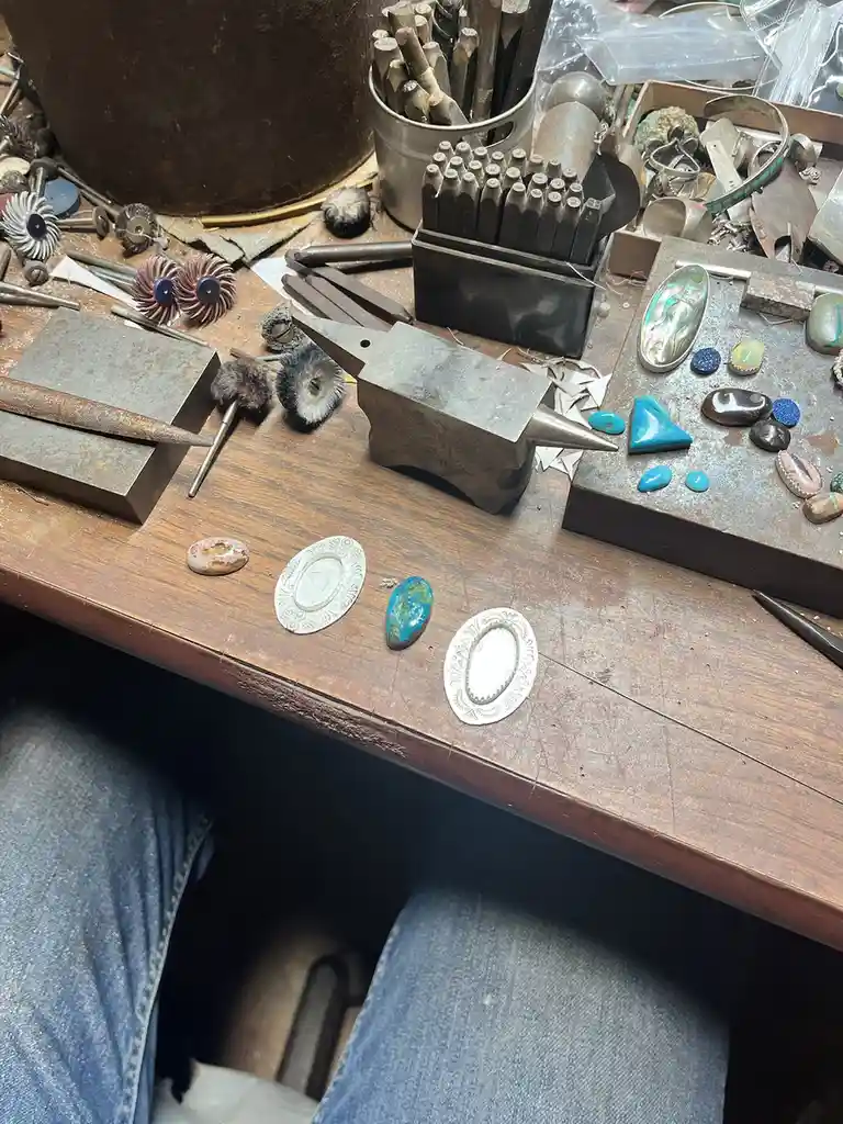 A cluttered jeweler's workbench is filled with tools like an anvil, punches, brushes, and various gemstones including…