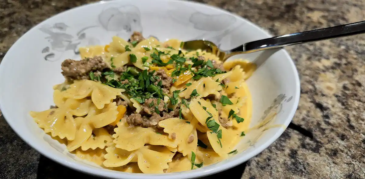 A close-up photo shows a white bowl with a grey floral pattern, filled with bow-tie pasta in a creamy sauce, mixed with…