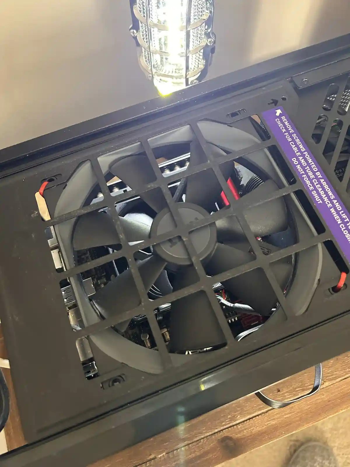 A close-up photo of a computer case's internal fan, viewed through a black plastic grid. A purple sticker with white text is…