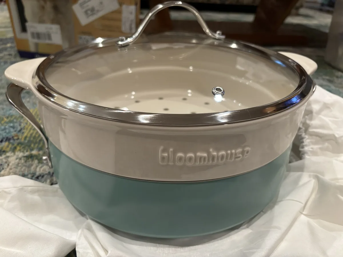 A two-toned ceramic pot with a glass lid and stainless steel handles, branded 'bloomhouse', sits on a white cloth. The pot…