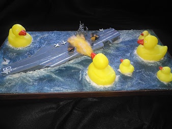 A cake shaped like an aircraft carrier is surrounded by several yellow rubber ducks floating on a blue frosting ocean. One…