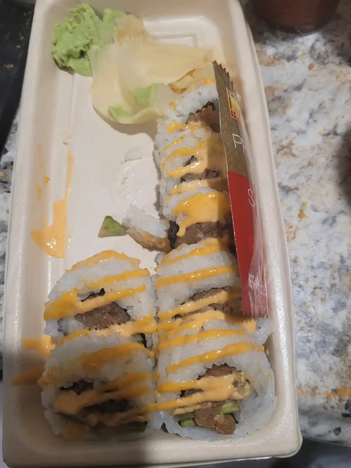 A takeout container holds sushi rolls drizzled with a thick orange sauce, with a small portion of wasabi and pickled ginger…