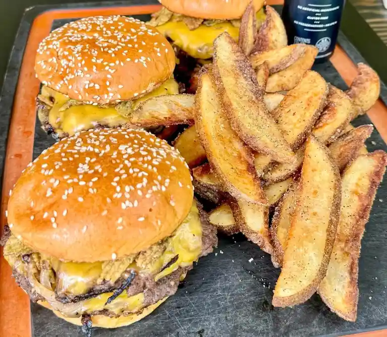 A close-up photo shows two large cheeseburgers with sesame seed buns and a generous side of seasoned potato wedges on a…