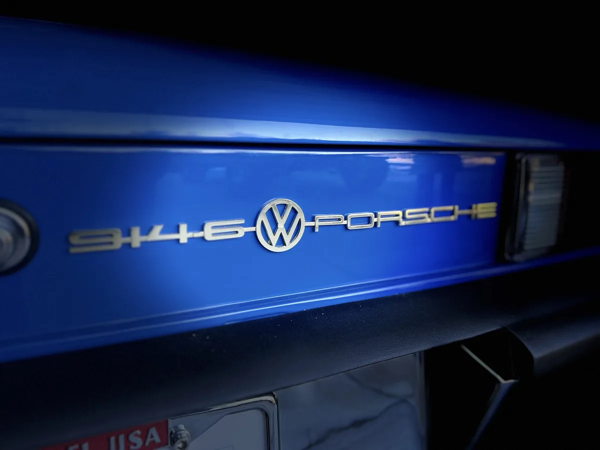 A close-up of a blue car's rear badge shows the number '946' followed by a Volkswagen logo and the word 'PORSCHE' in silver…