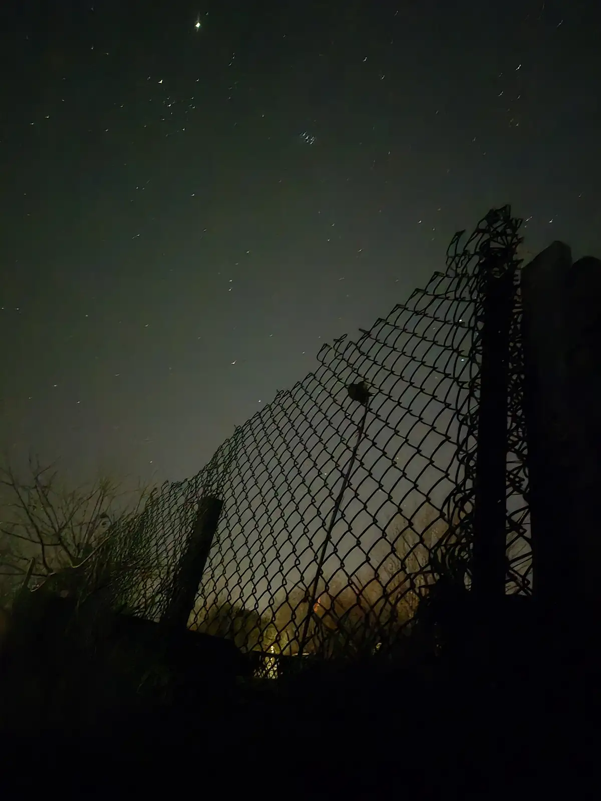 A low-angle nighttime photo shows a chain-link fence silhouetted against a dark sky filled with stars and faint light…