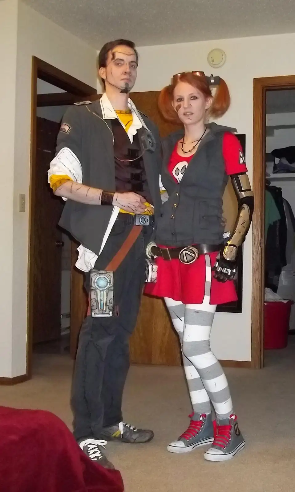 Two people dressed as characters from the video game Portal, standing indoors. The man on the left wears a grey vest over a…