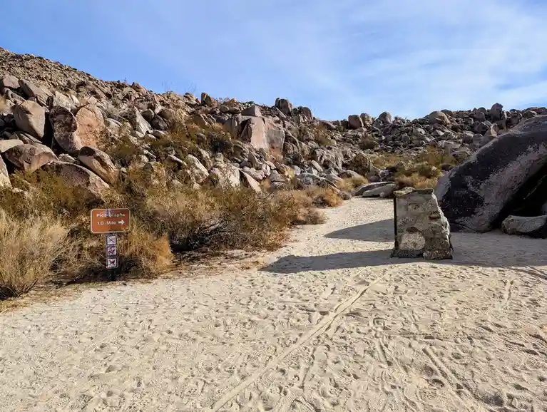 A desert trail with a brown sign indicating 'Pictographs 1.0 Mile →' and three small prohibitive symbols below it, set…