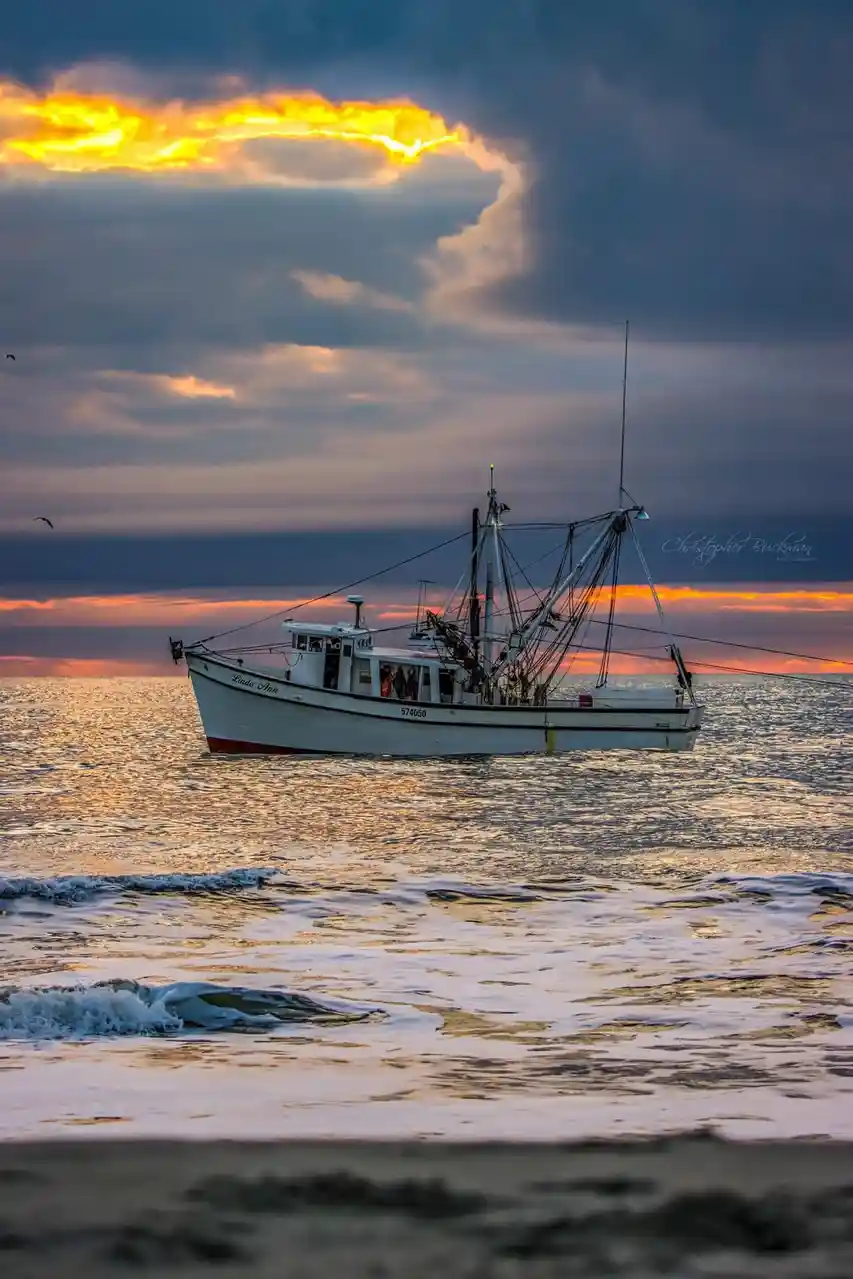 A white fishing boat named 'Linda Ann' with hull number 574050 floats calmly on the ocean at sunset, its silhouette framed…