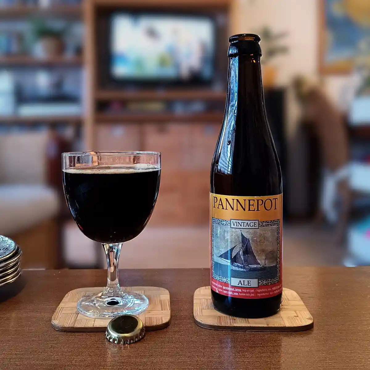 A dark bottle of 'Pannepot Vintage Ale' beer sits next to a filled glass on wooden coasters, with a bottle cap nearby on a…