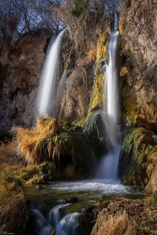 A long-exposure photograph of a natural waterfall cascading down mossy, rocky cliffs into a stream below, with bare trees…