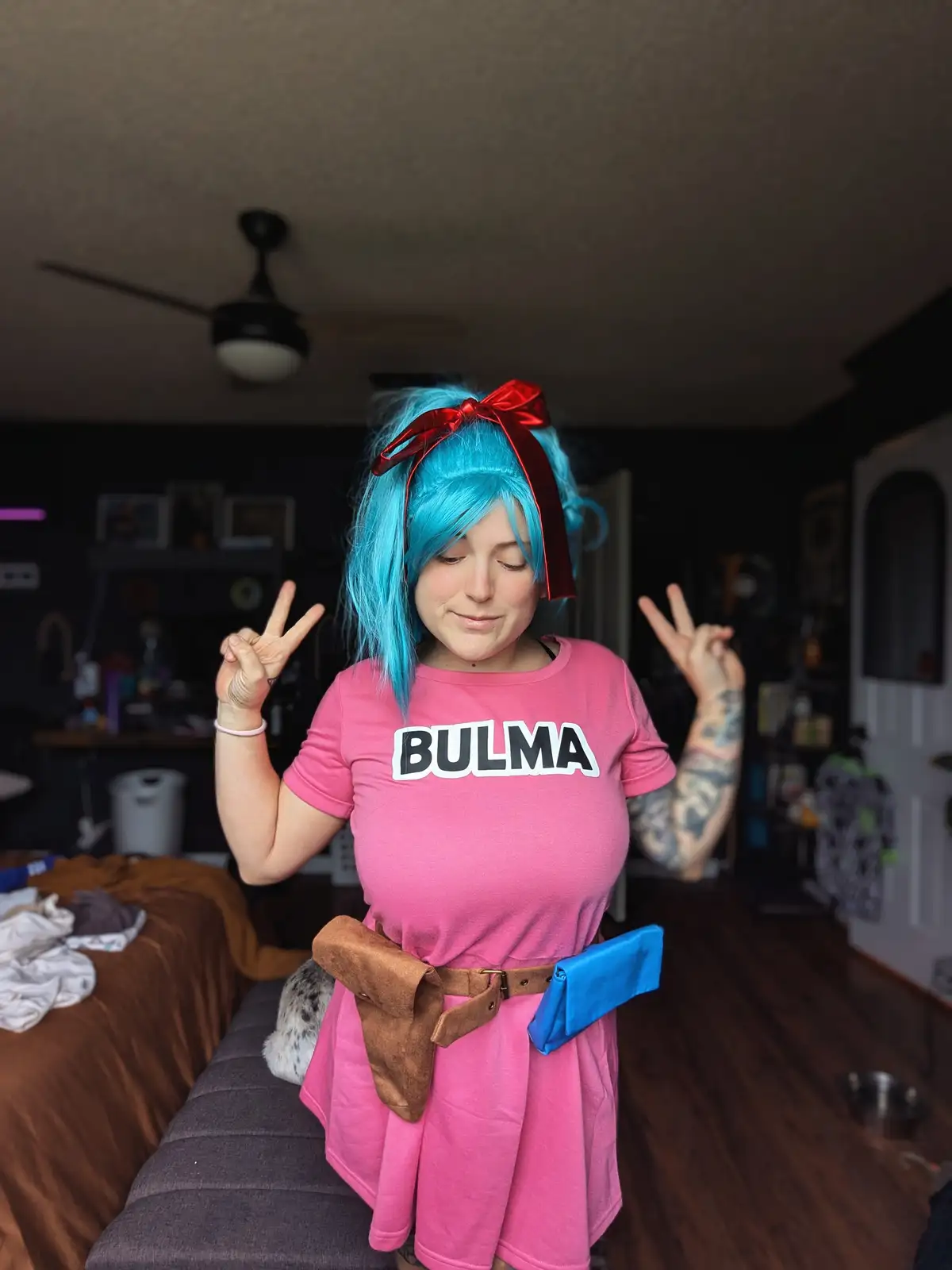 A person with blue hair tied with a red bow, wearing a pink shirt with 'BULMA' printed on it, stands indoors making peace…