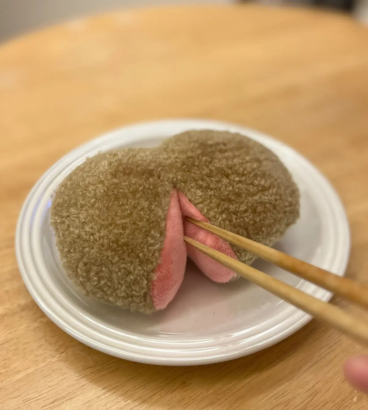 a close-up photo shows a brown, textured, bunny-shaped mochi dessert on a white plate, with wooden chopsticks gently poking…