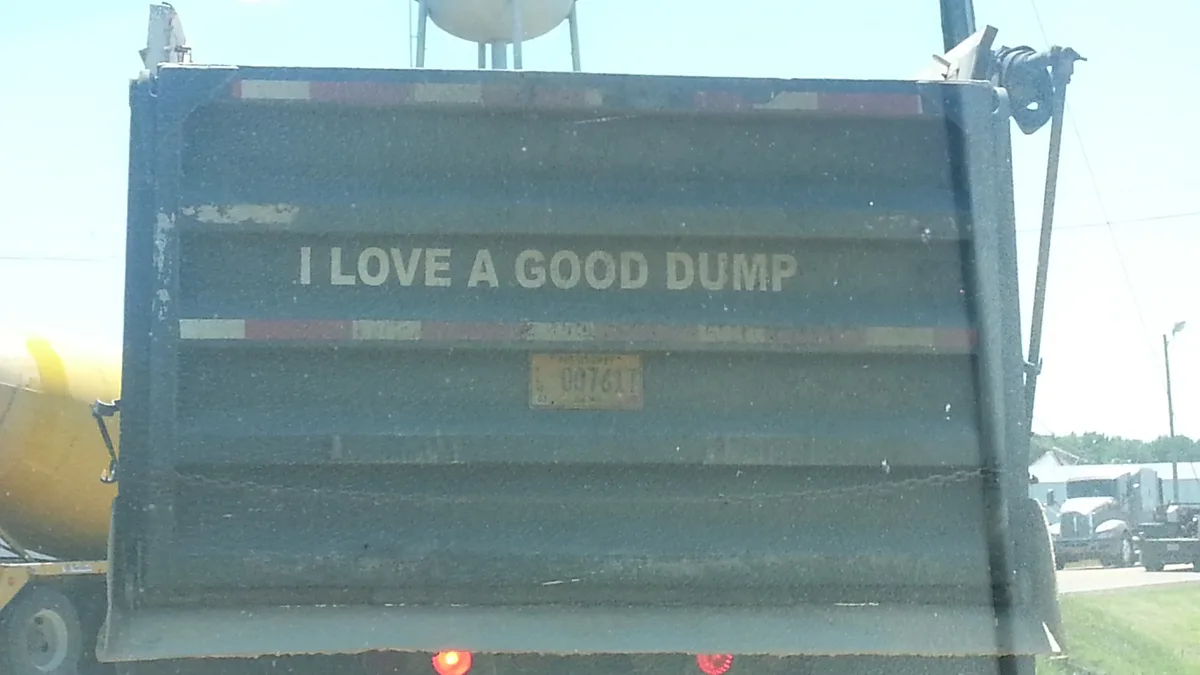 The image shows the rear of a large, dark green dump truck with the phrase 'I LOVE A GOOD DUMP' painted in white capital…