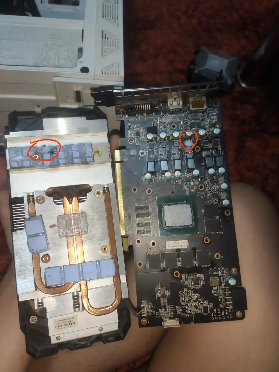 A person holds a disassembled graphics card and its attached cooler side-by-side, showing visible damage. Red circles…