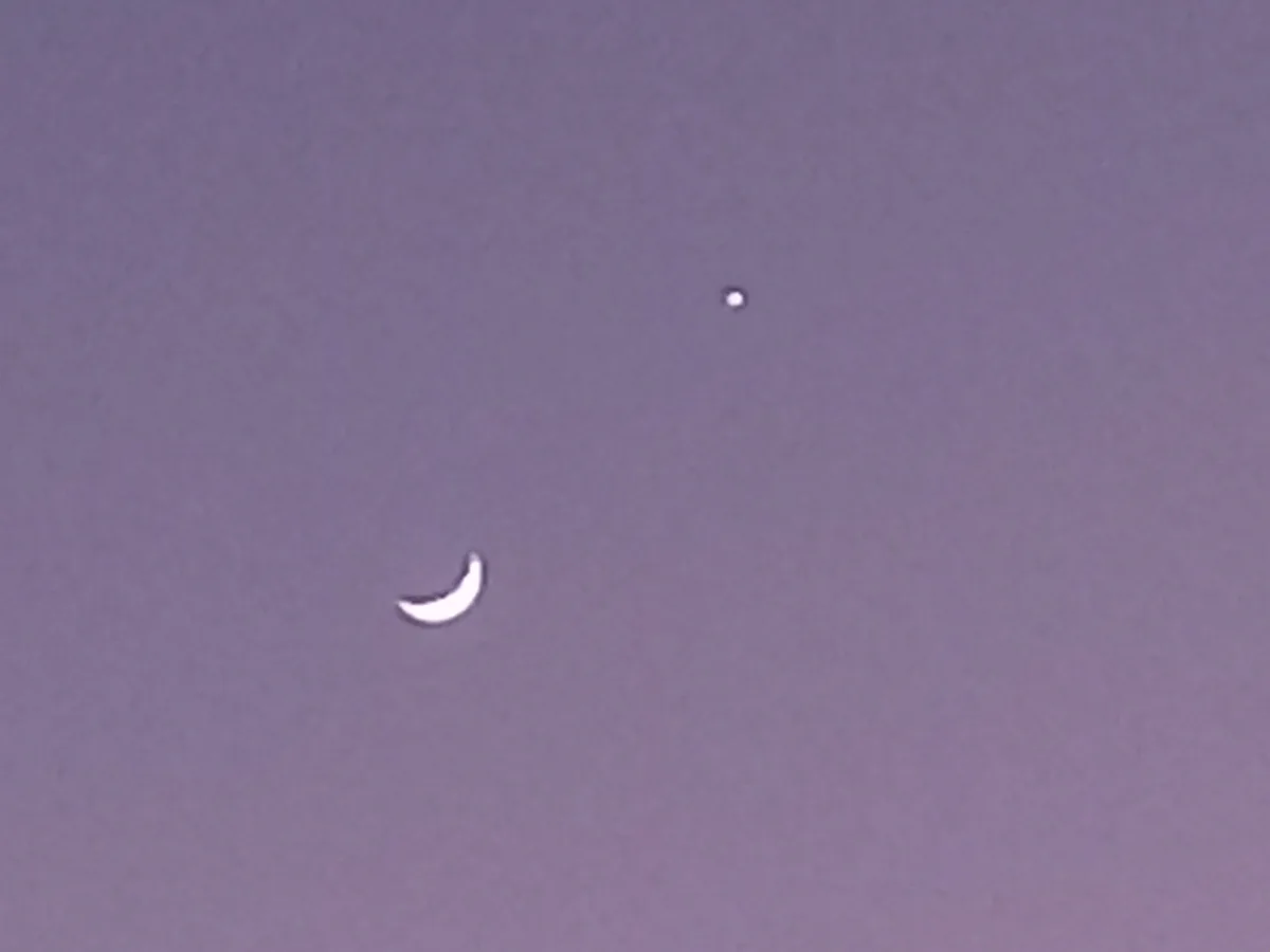 A pale purple sky shows a crescent moon and a small bright dot, likely a planet or star, positioned near the moon. The image…