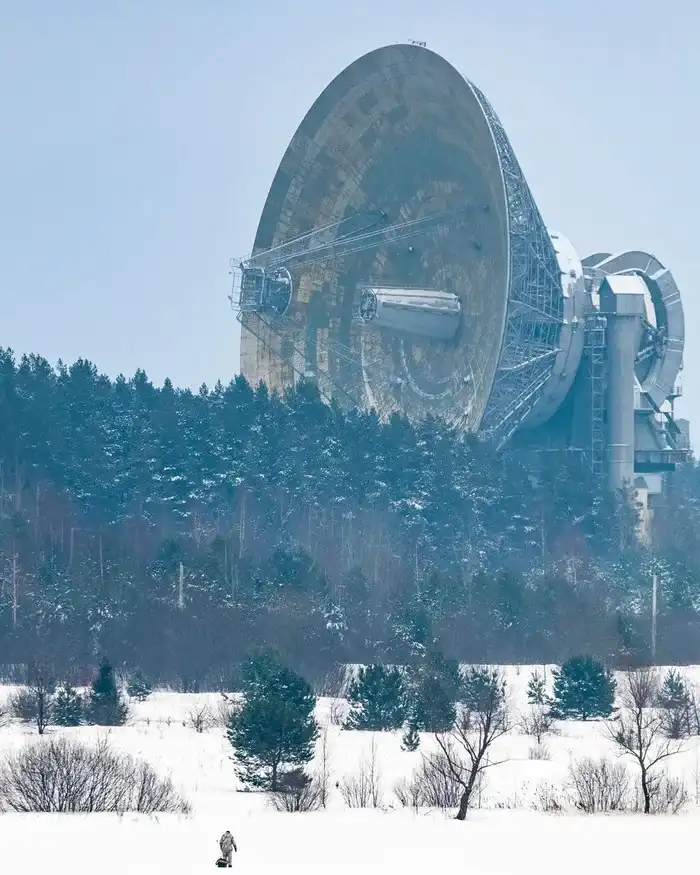 A tiny person in a winter coat walks across a snowy field toward a massive radio telescope, with a forest behind them under…