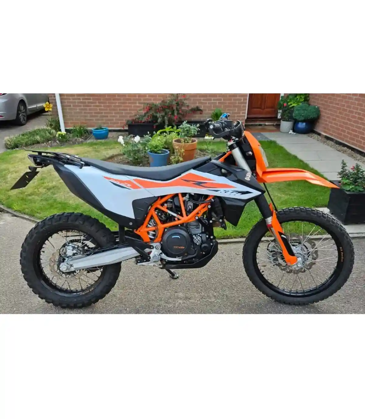 A side-profile photo of an orange and white KTM dirt bike parked on a driveway in front of a brick house with potted plants…