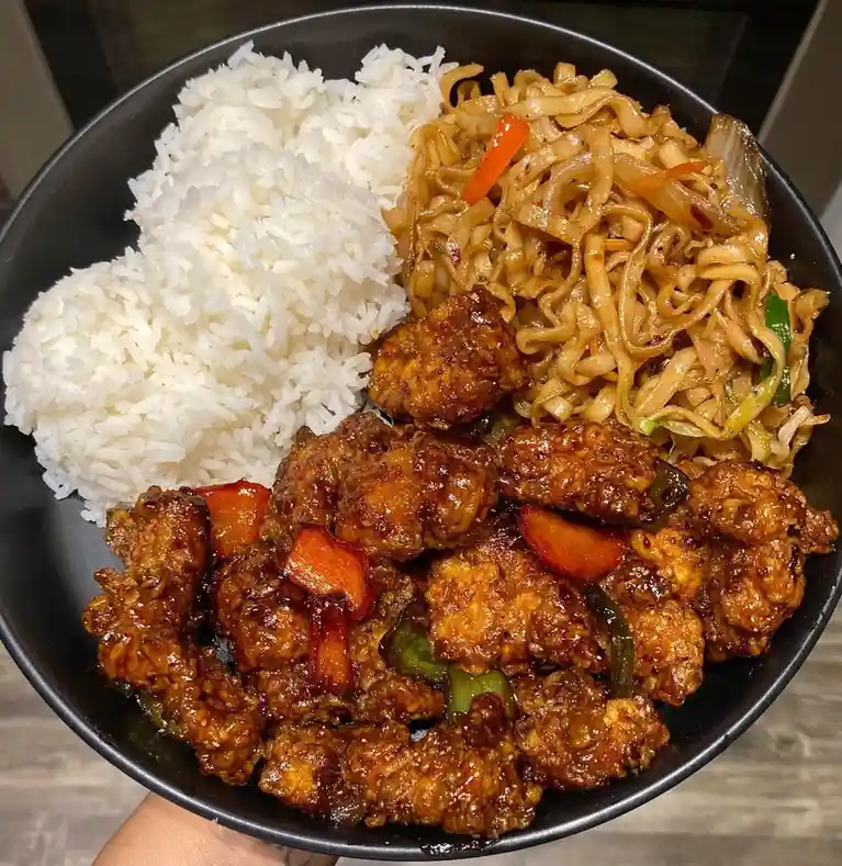 A close-up photo of a black bowl filled with a meal consisting of white rice, stir-fried noodles with vegetables, and…