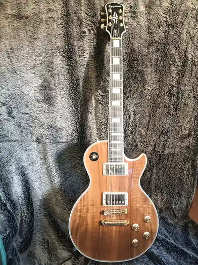 A light brown Epiphone Les Paul Custom Pro electric guitar with gold hardware and white block inlays rests vertically on a…
