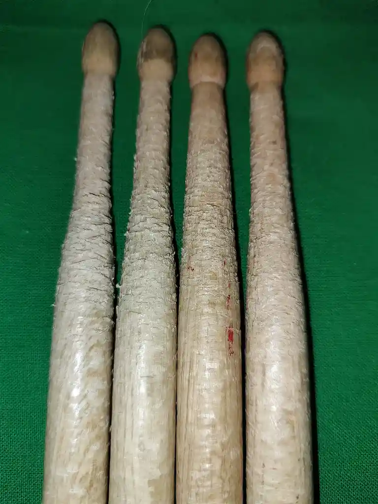 Four worn wooden drumsticks are lined up vertically on a green fabric background. The sticks show significant use, with…