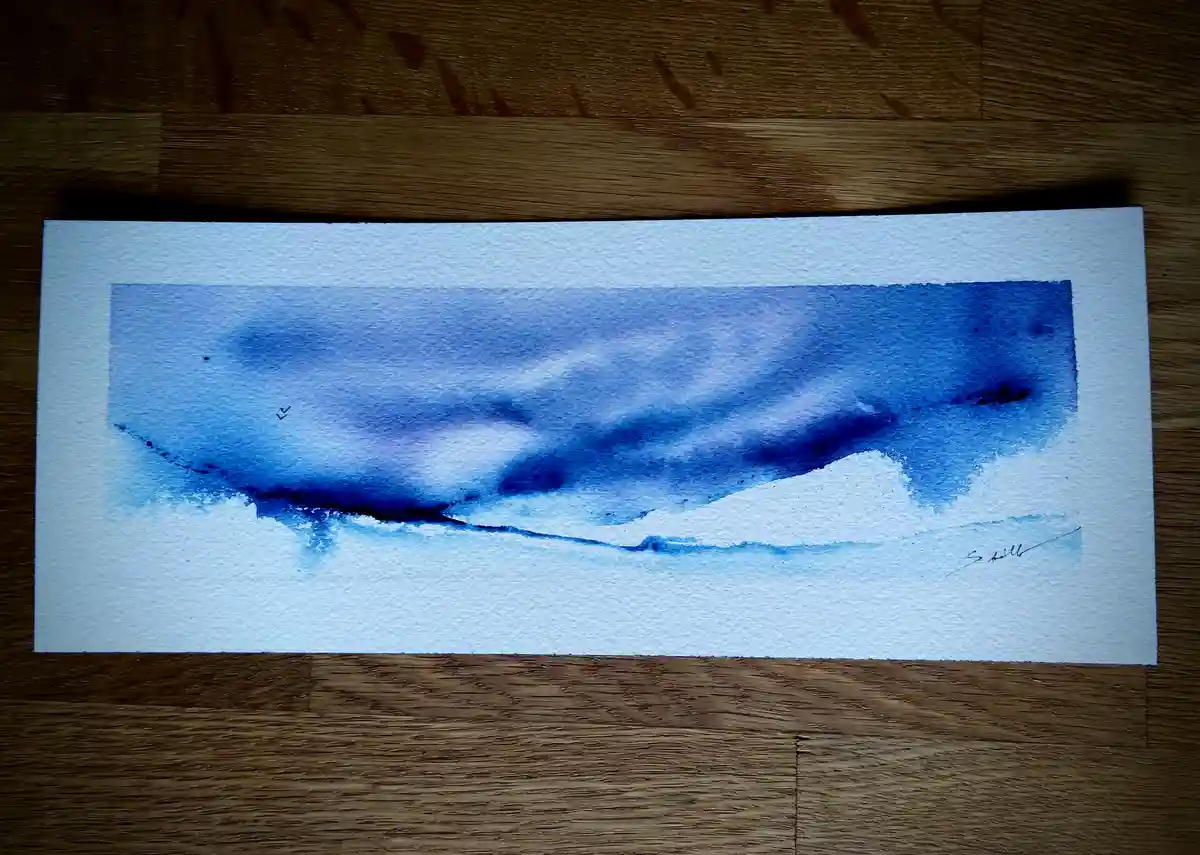 A watercolor painting on white paper, resting on a wooden surface, depicts a large blue whale swimming underwater with a…