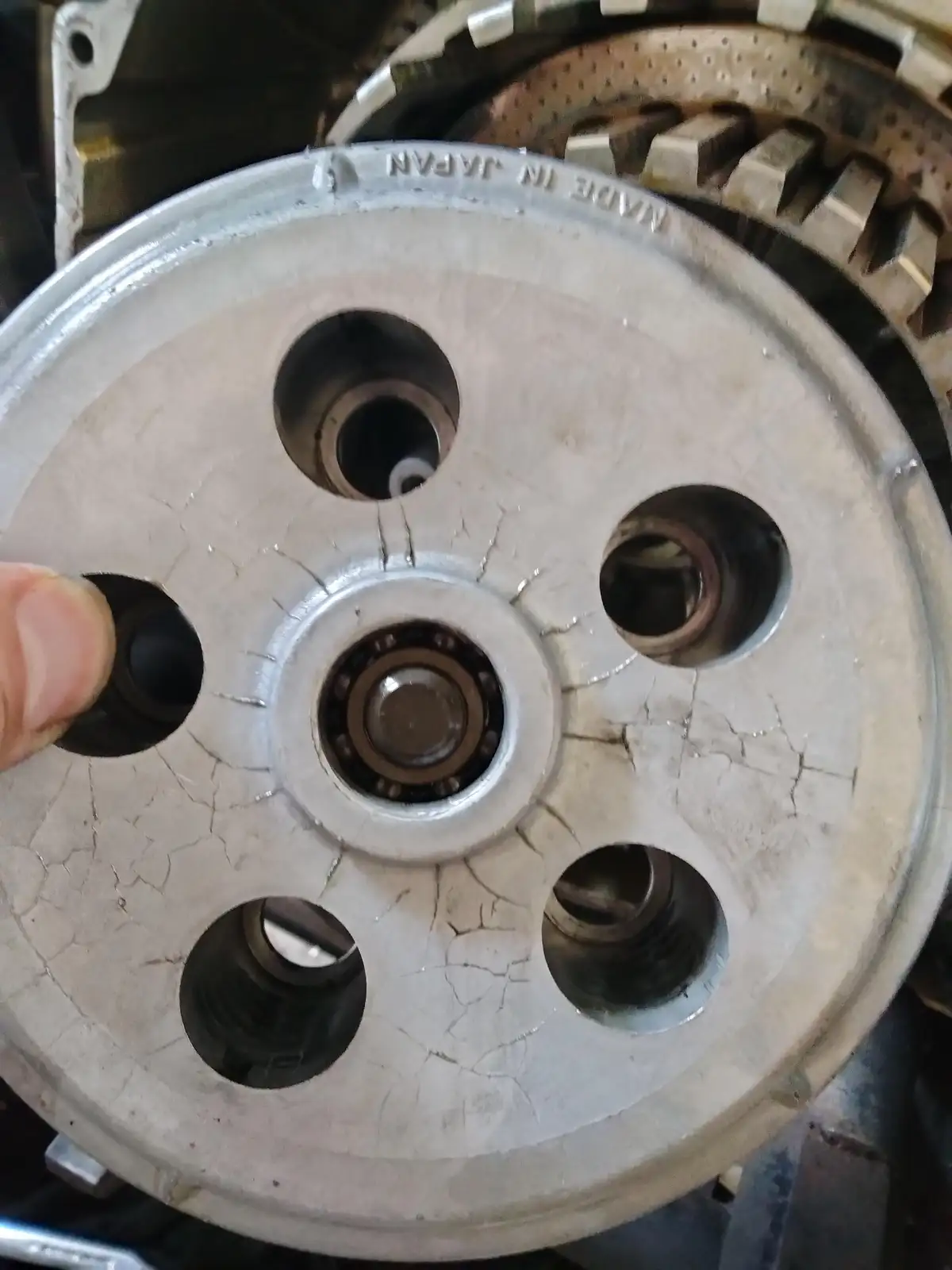 A close-up photo shows a person's thumb holding a cracked, silver-colored metal wheel hub with five bolt holes and a central…