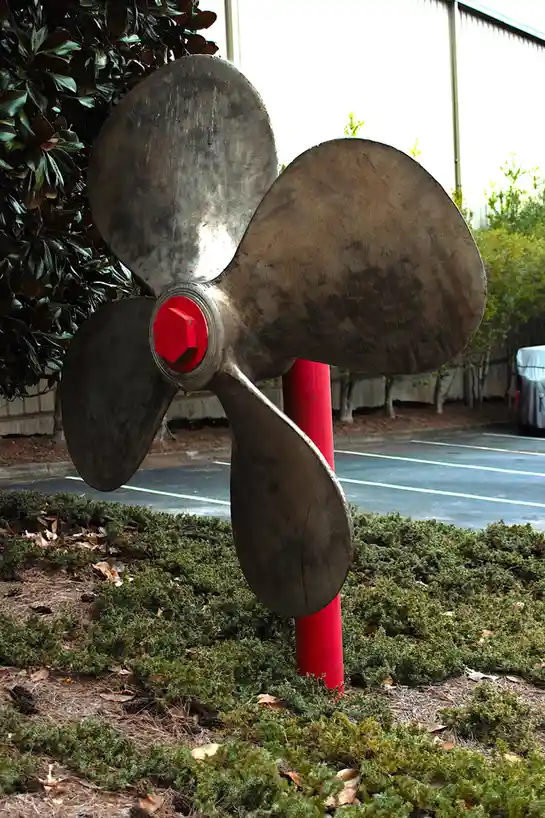 A large, three-bladed metal boat propeller with a red central hub and bolt is mounted vertically on a red pole, standing in…